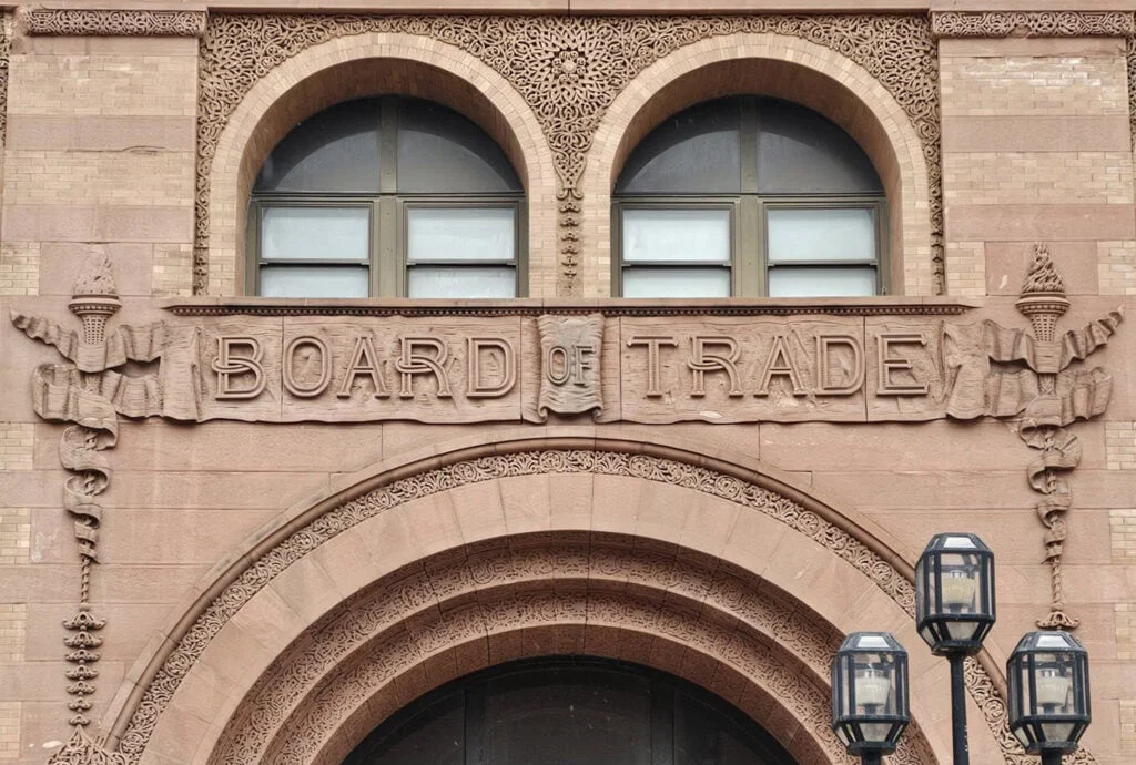 A close-up of an ornate stone building facade with two arched windows above a carved sign that reads "BOARD OF TRADE." Decorative columns and torches frame the detailed inscription. Three modern street lamps are in front.