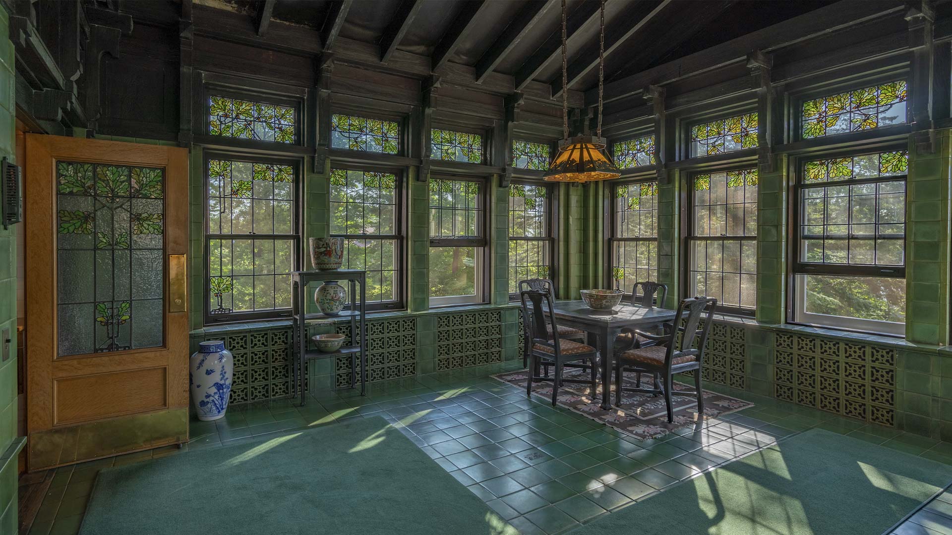 The Cotton Mansion A sunlit room with green tiled floors and walls, stained glass windows, a wooden table with four chairs, a hanging stained glass lamp, and decorative pottery accents.