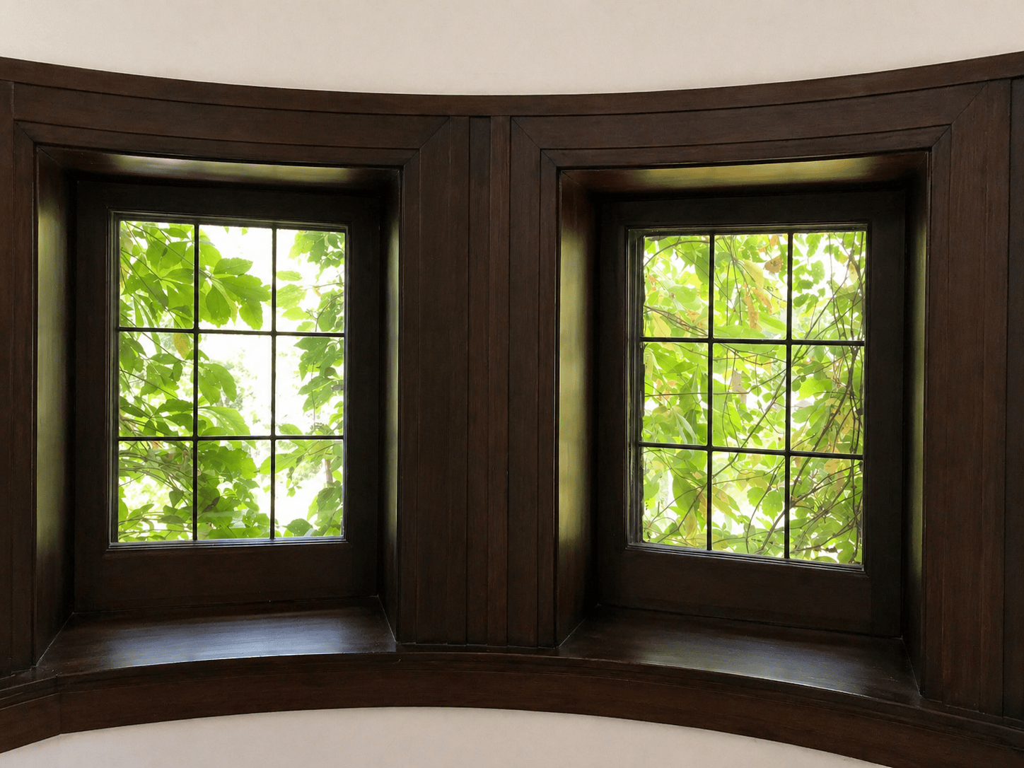 Two wooden-framed windows with grid panes set in a curved wall, showing lush green leaves and branches outside, allowing natural light to enter the interior space.