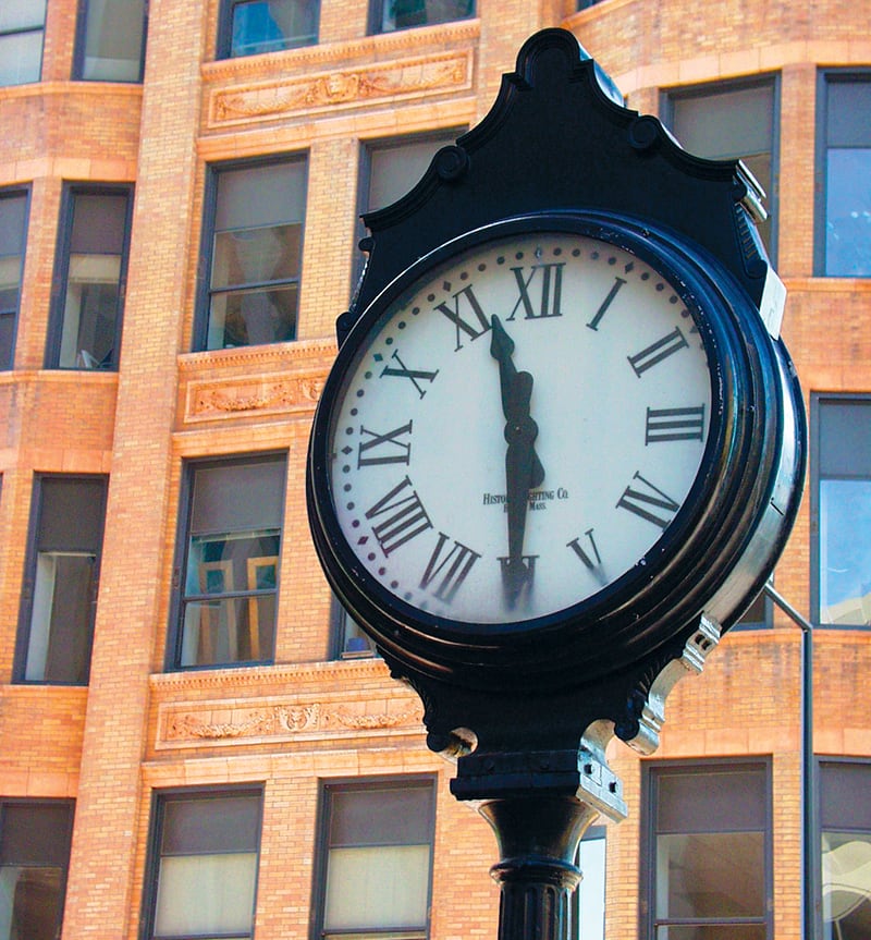 A large black street clock with Roman numerals shows the time as 12:00, standing in front of a tan brick building with many windows.