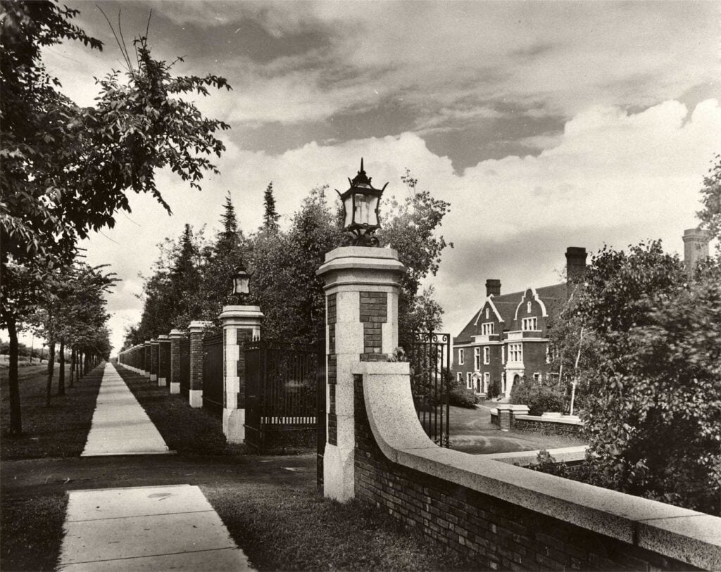 Black-and-white photo of a long brick and iron fence with lanterns, running alongside a tree-lined sidewalk. In the background, a large, stately house with peaked roofs is partially visible among trees.