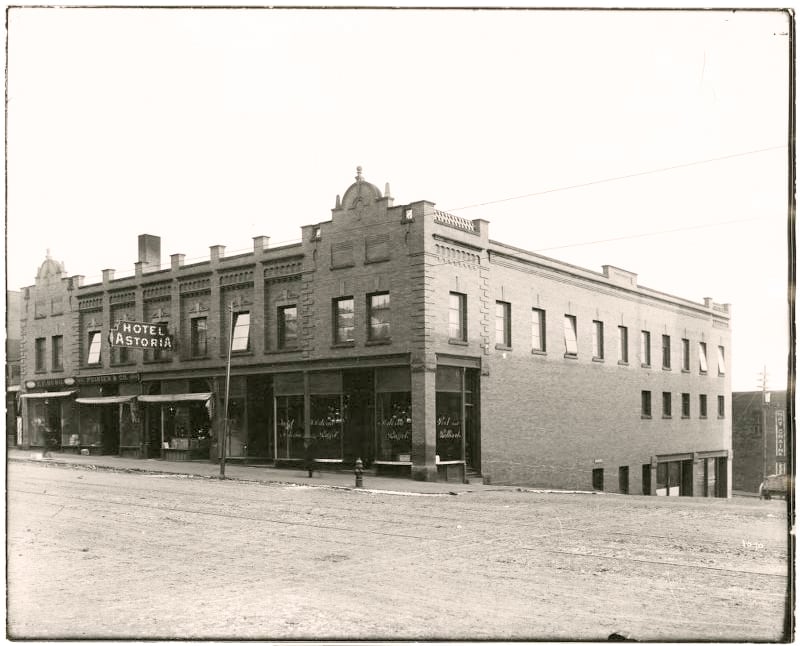 Black-and-white photo of a two-story brick building on a dirt street corner, featuring the "Hotel Astoria" sign and large storefront windows on the ground floor.