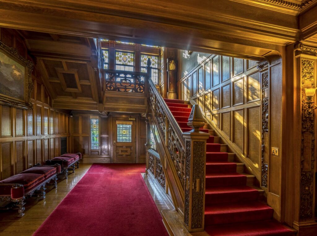 Ornate wooden staircase with red carpet runner in a grand hallway, stained glass windows, carved wood paneling, benches with red cushions, and soft sunlight streaming through the windows.
