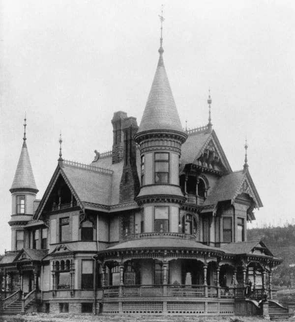 Black and white photo of a large, ornate Victorian house with multiple pointed towers, intricate woodwork, and a wraparound porch, showcasing elaborate architectural details.