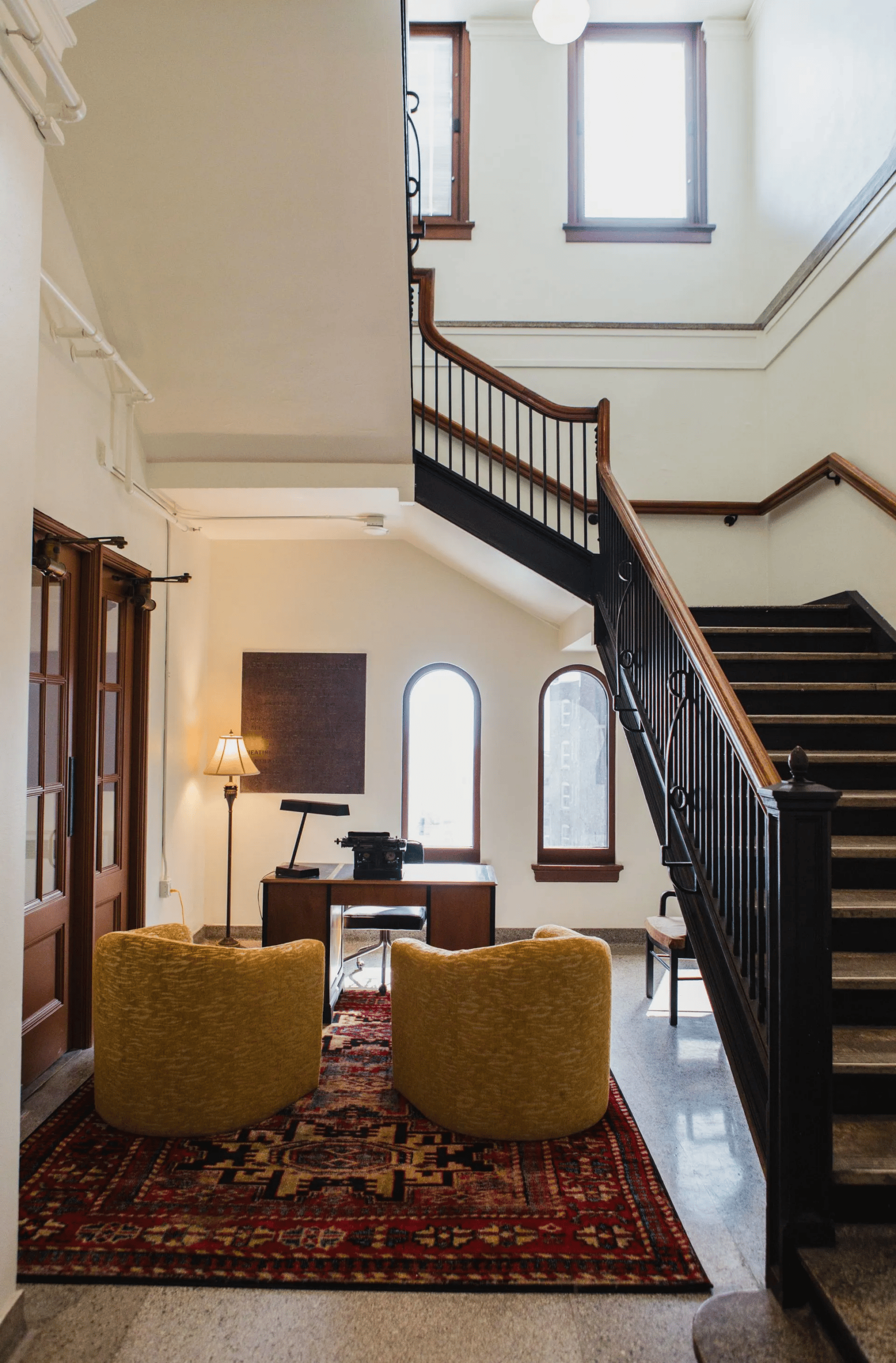 Zenith Apartments A cozy office space with two yellow chairs on a patterned rug faces a wooden desk under a staircase. A lamp and large windows provide natural light. The stair railing is black with wooden accents.