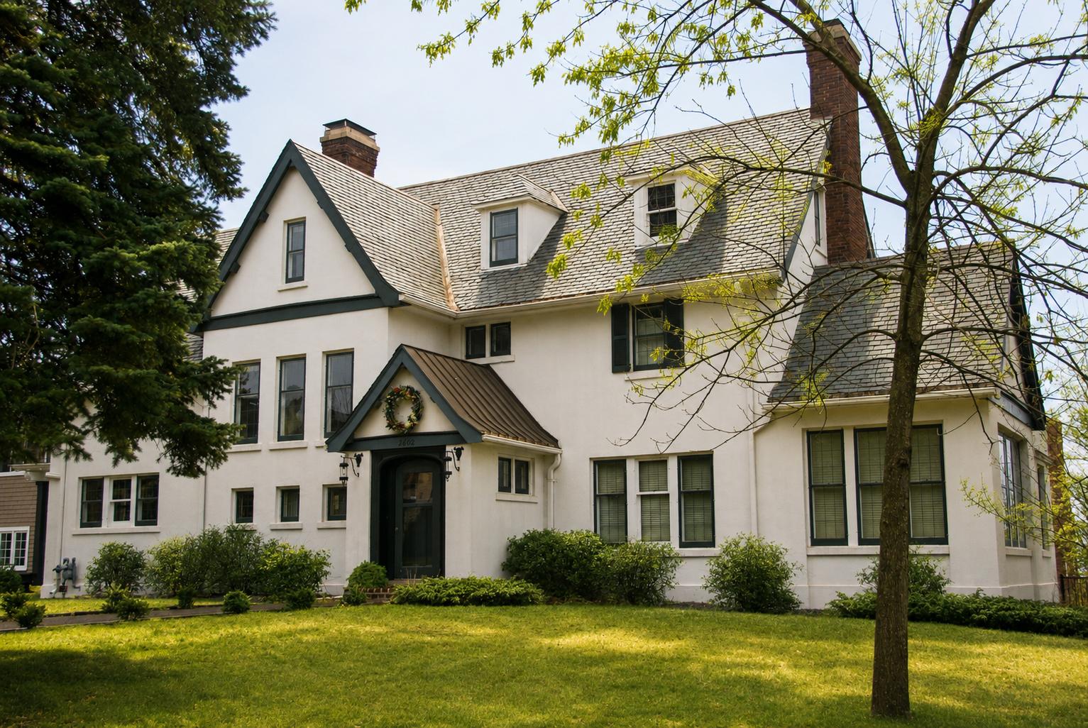 A large white two-story house with dark trim, a steep roof, multiple windows, and a front porch, surrounded by green grass, shrubs, and trees on a sunny day.