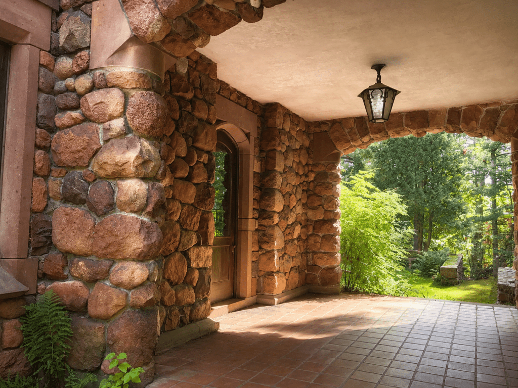 A covered porch with stone walls and arches, tiled floor, and a lantern hanging from the ceiling, opens to a lush green garden with trees and shrubs in the background.