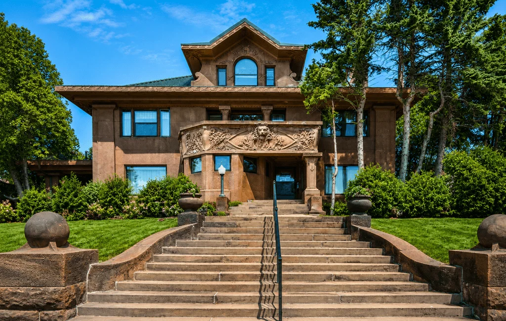 A large historic mansion with ornate stonework, arched windows, and a grand staircase leading to the entrance, surrounded by green lawns, shrubs, and tall trees under a blue sky.