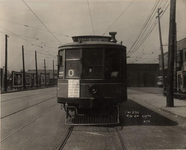 A vintage streetcar sits on tracks in an empty street, facing the camera. Overhead wires, utility poles, and old buildings line the street. A sign on the streetcar reads "CAR HOUSE ONLY.