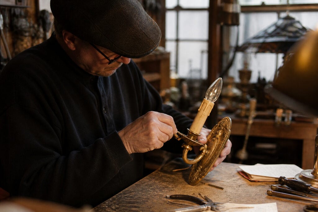 A person wearing a flat cap and glasses repairs a vintage brass wall sconce with a candle-shaped bulb at a wooden workbench in a workshop filled with tools and antique objects.