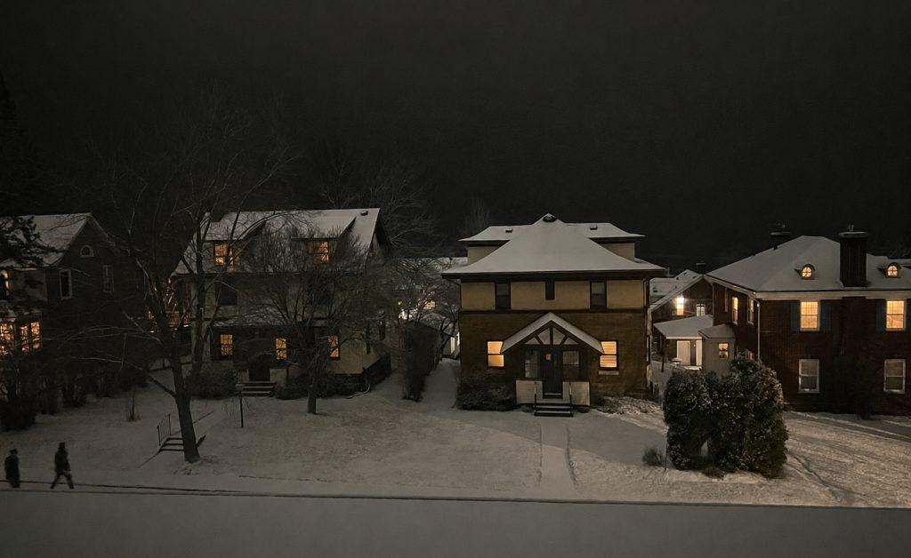 A nighttime scene shows snow-covered houses with lights glowing from the windows. Two people walk along a sidewalk lined with bare trees. The sky is dark, and the ground and rooftops are blanketed in snow.