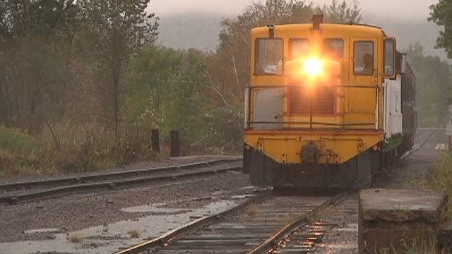 A yellow train engine with its headlight on sits on wet railroad tracks surrounded by trees and mist in a rural area.