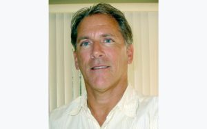 A middle-aged man with short brown hair, wearing a light-colored collared shirt, stands in front of vertical blinds, looking at the camera with a neutral expression.