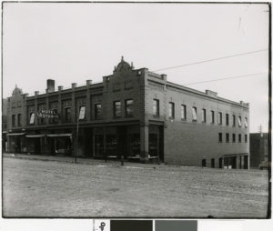 Black-and-white photo of a two-story brick building on a corner, with a sign reading “Hotel Astoria.” The street in front is unpaved and there are large windows on both floors.