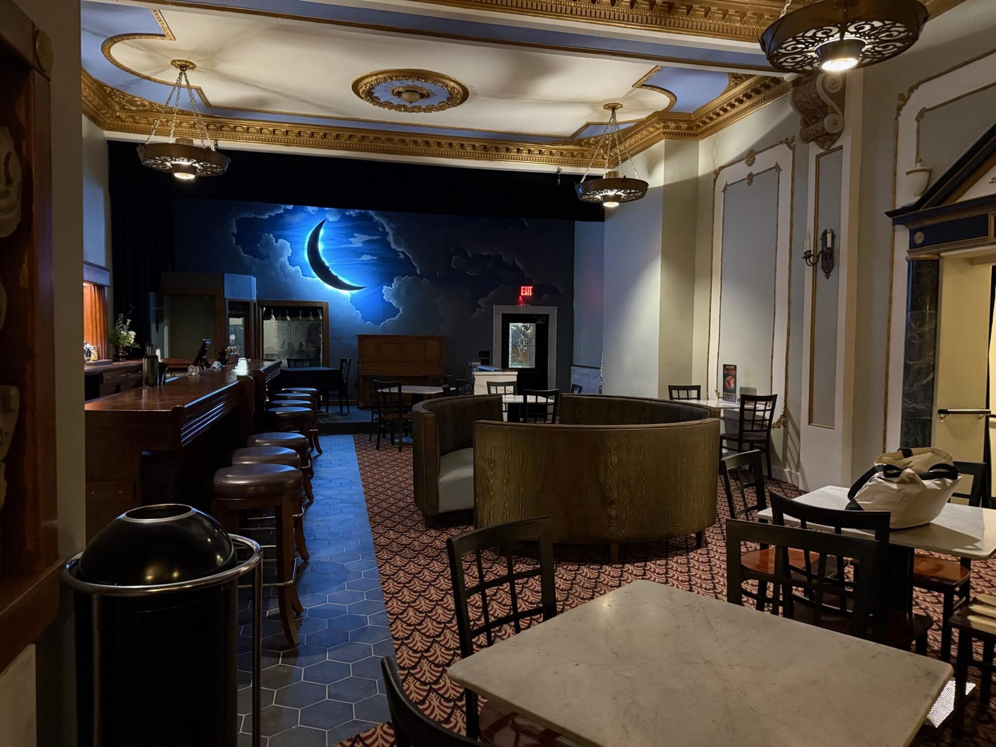 A dimly lit cafe or bar with round and square tables, wooden chairs, a bar on the left, and a stage at the back featuring a glowing crescent moon and clouds on a dark blue backdrop. Ornate ceiling and wall details are visible.