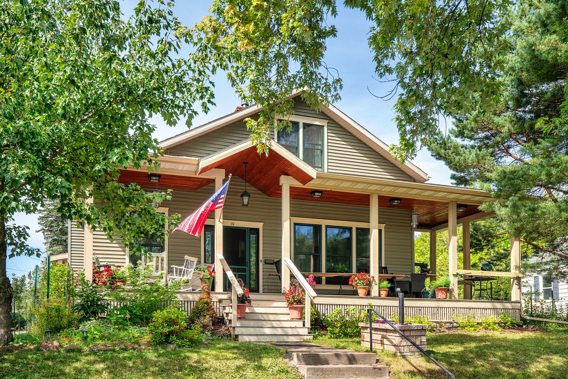 A cozy beige house with a front porch, American flag, potted plants, and outdoor seating, surrounded by lush greenery and trees on a sunny day. Steps lead up to the porch from a landscaped yard.