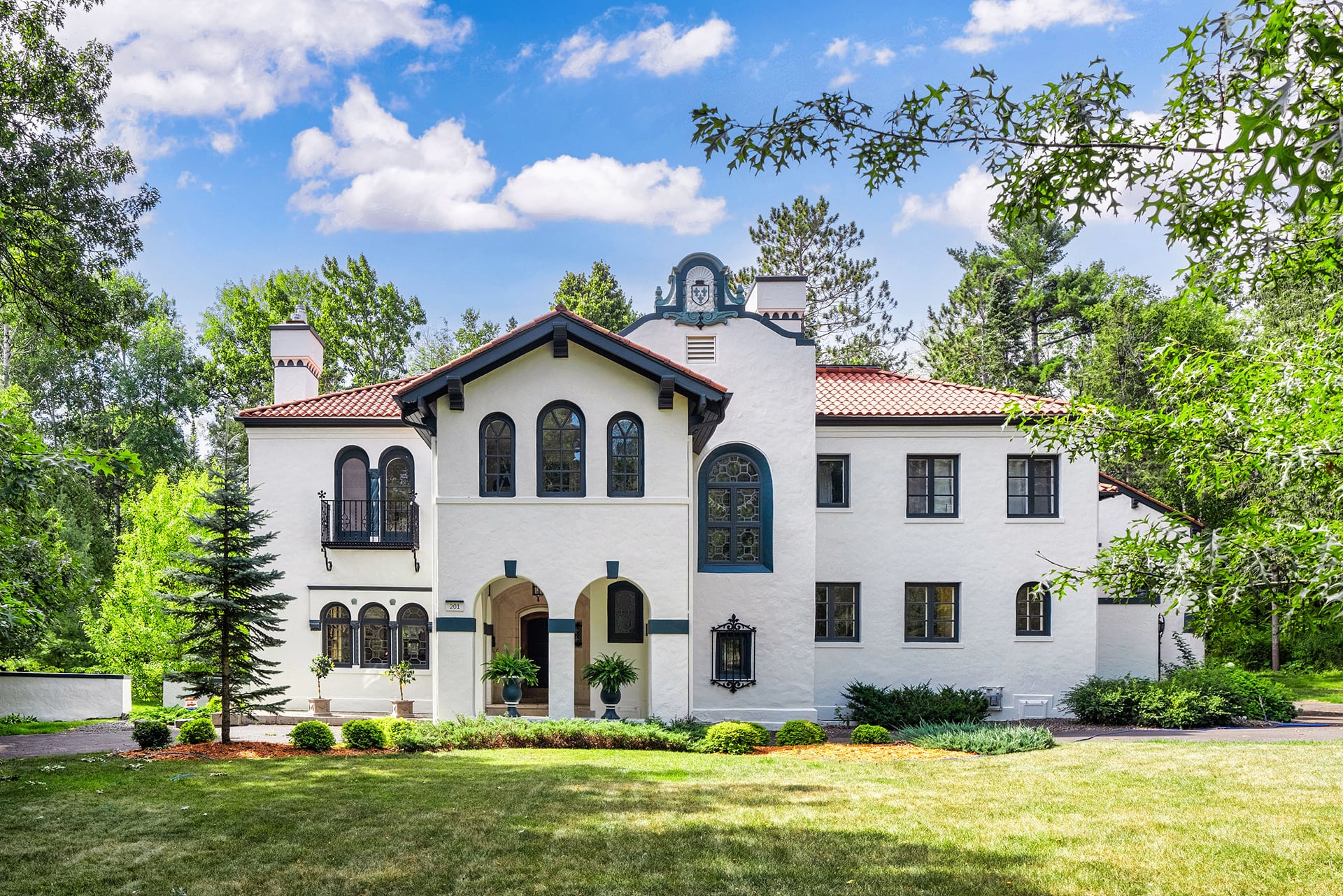 A large two-story white stucco house with a red tile roof, arched windows and doorways, surrounded by green trees and a manicured lawn, under a blue sky with scattered clouds.