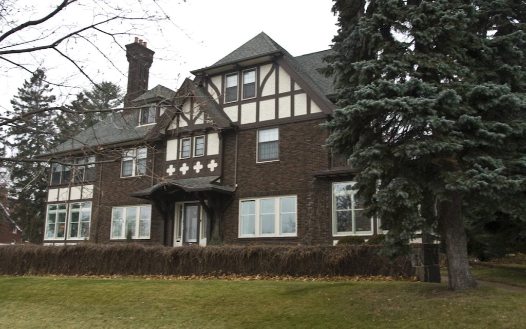 Large historic Tudor-style house with dark brick and white accents, peaked roofs, prominent chimneys, and many windows, surrounded by green lawn, brown hedges, and tall evergreen trees on a cloudy day.