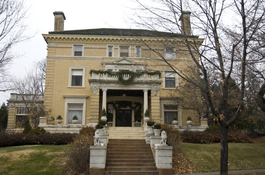 A large, elegant, beige mansion with tall columns, green-tiled roof, and stone steps leading to the entrance, flanked by two white lion statues and surrounded by leafless trees and landscaped bushes.