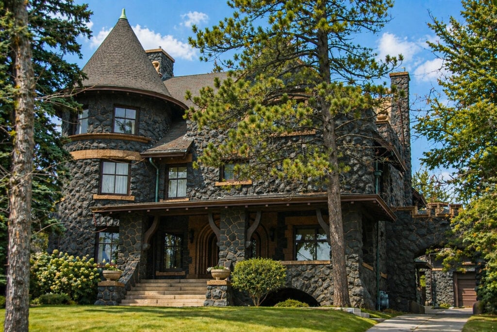 A large, historic stone mansion with a turret, arched porch, and steep roof is surrounded by trees and greenery on a sunny day.