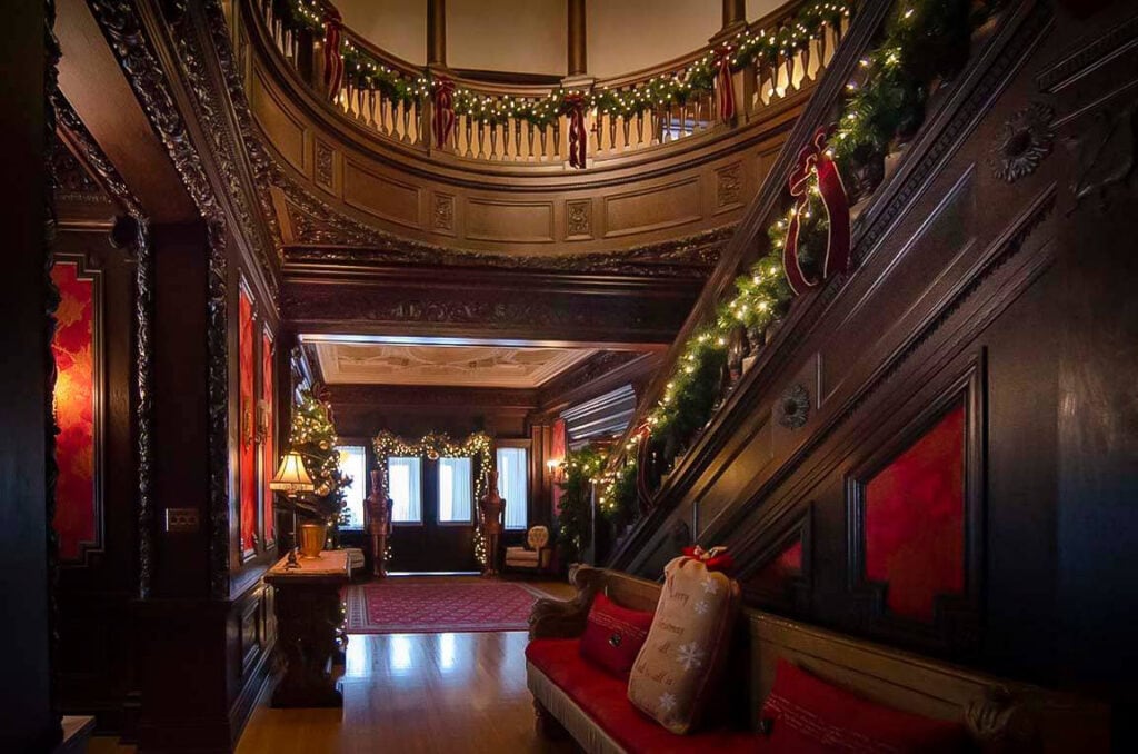 Grand wooden staircase and balcony decorated with garlands and Christmas lights, leading to an ornately detailed upper level. A cozy seating area with red cushions sits by the stairs in the warmly lit, festive interior.
