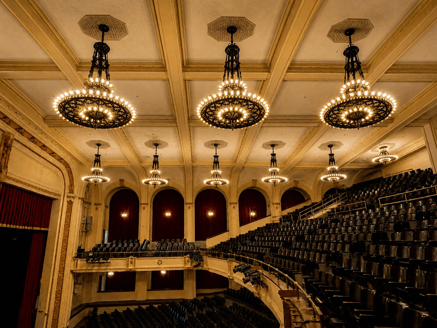 An elegant theater interior with ornate chandeliers hanging from the ceiling, rows of empty dark seats, red-arched curtains along the walls, and a stage with a closed red curtain on the left.
