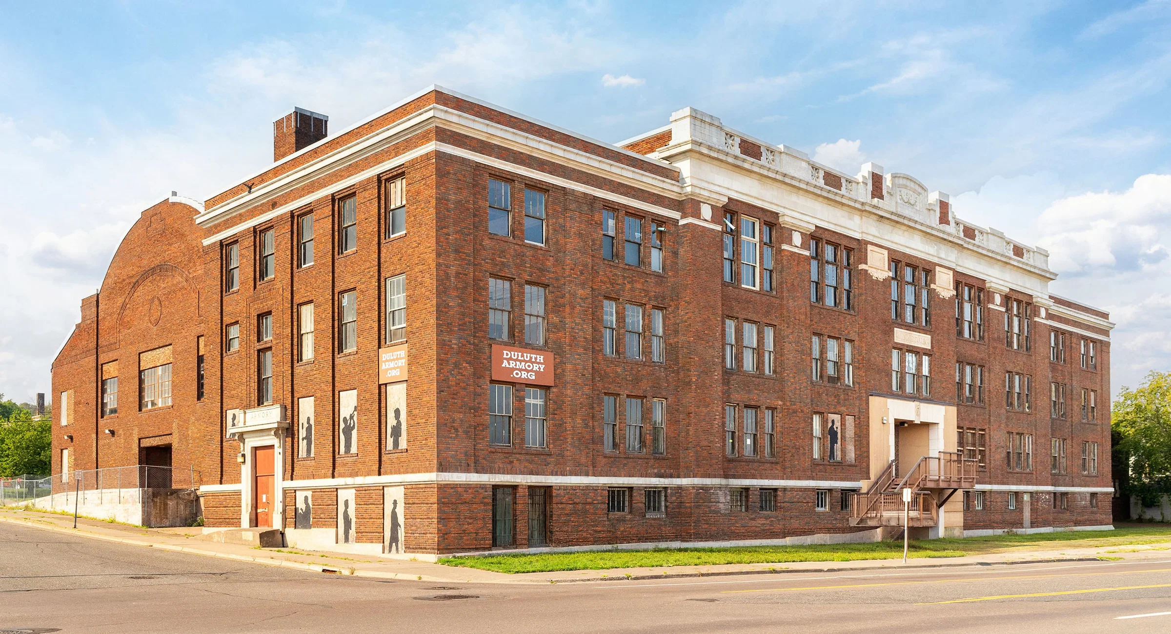 A large, historic red brick building with many windows and a small staircase at the entrance, seen from the corner of a quiet street under a blue sky with clouds.
