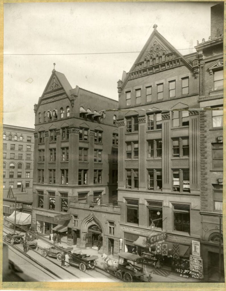 Historic black-and-white photo of a busy city street lined with ornate, multi-story buildings featuring shops on the ground floor and parked early 20th-century automobiles and wagons along the curb.