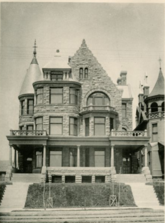 Black-and-white photo of a large, ornate Victorian-style stone mansion with turrets, arched windows, and steeply pitched roofs, set on a raised lot with steps leading to the front entrance.
