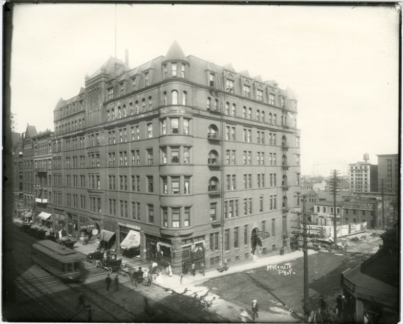 Black and white photo of a large, multi-story brick hotel on a city corner, with people, horse-drawn carriages, and a streetcar in front. Early 20th-century architecture and bustling street scene are visible.
