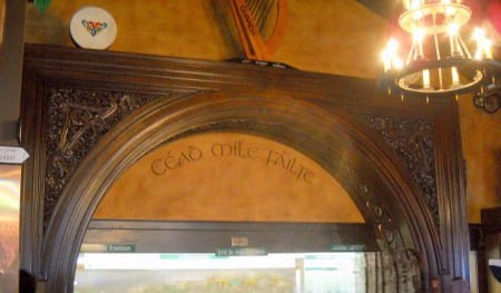 A wooden archway with the Irish phrase “Céad Míle Fáilte” carved above it. Decorative carvings and a harp are above the arch, with a chandelier hanging to the right.
