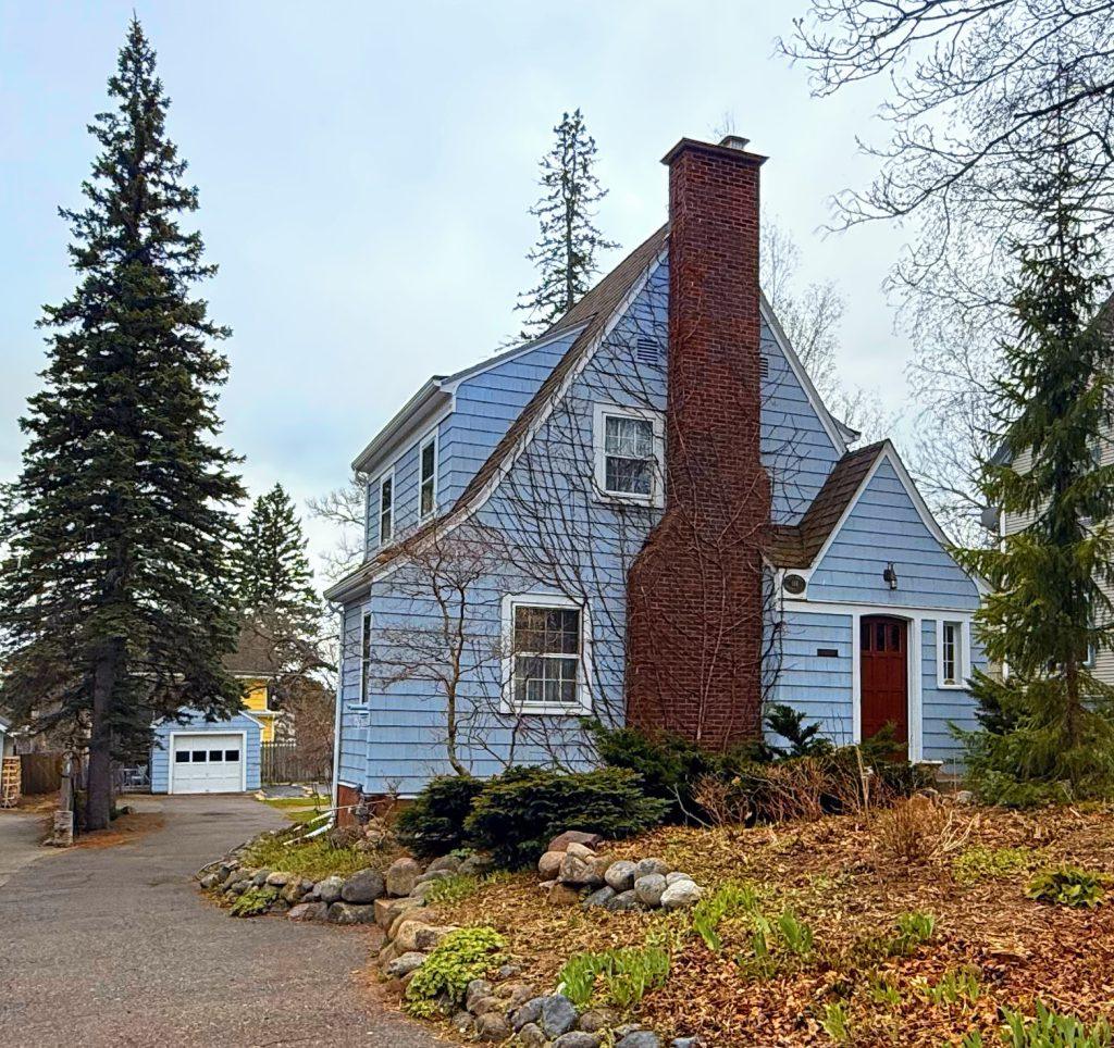 A blue two-story house with white trim and a red front door, featuring a tall brick chimney. The yard has rocks, bare shrubs, and evergreen trees, with a driveway leading to a detached white garage.