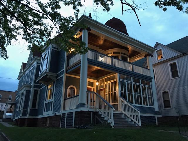 A two-story Victorian-style house with blue siding, white trim, a wraparound porch, large bay windows, and a turret, lit warmly at dusk under a partly cloudy sky.