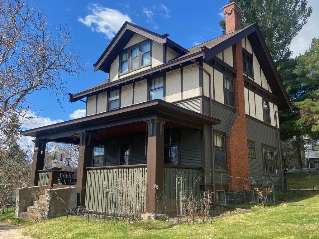 A two-story house with Tudor-style architecture, brown wood trim, a large brick chimney, and a covered front porch, set on a grassy lawn under a bright blue sky with scattered clouds.