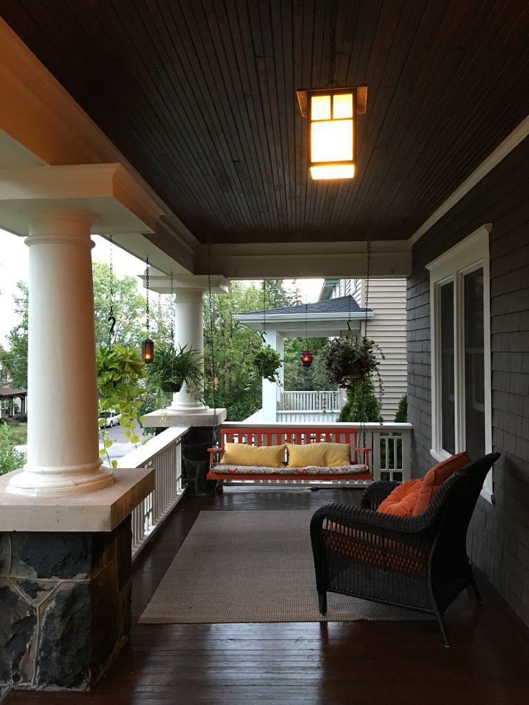 A cozy front porch with a wooden ceiling, large white columns, a hanging porch swing with yellow cushions, a wicker chair with orange pillows, hanging plants, and a glowing overhead light at dusk.
