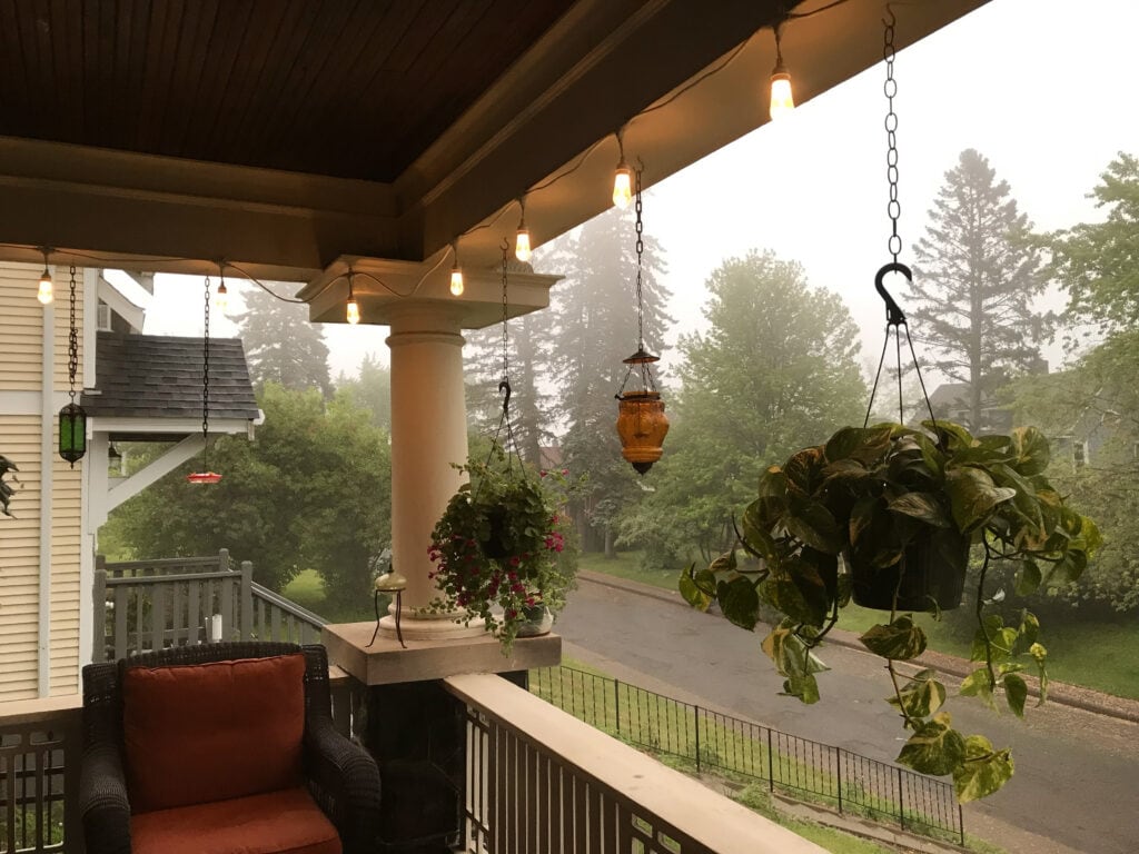 A cozy porch with hanging plants, a wicker chair with a red cushion, and string lights glowing in the foggy morning. Trees and a quiet street are visible in the background.