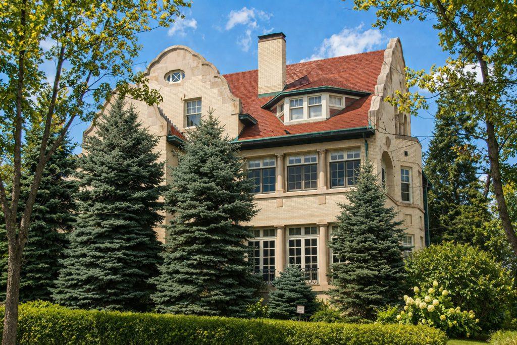 A large, beige brick house with a red roof and decorative gables, partially obscured by tall evergreen trees and surrounded by lush greenery under a blue sky with scattered clouds.
