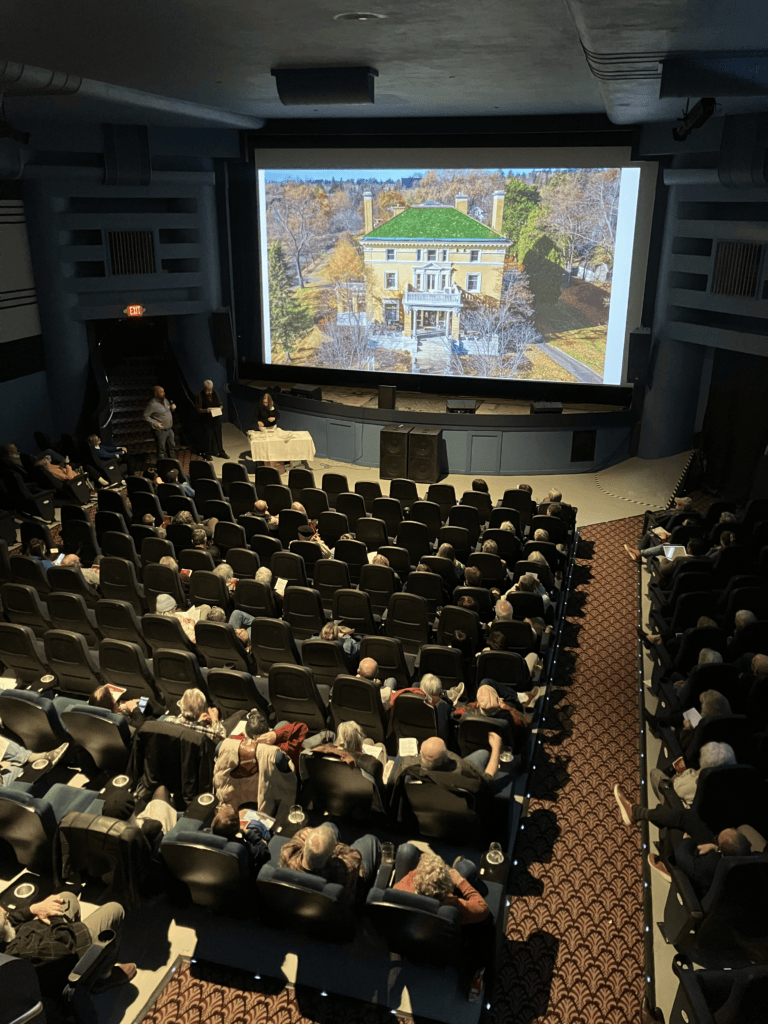 An audience sits in a theater watching a presentation. On the screen is an aerial photo of a mansion surrounded by autumn trees. Three presenters stand at the front near a table. The theater seats are mostly filled.
