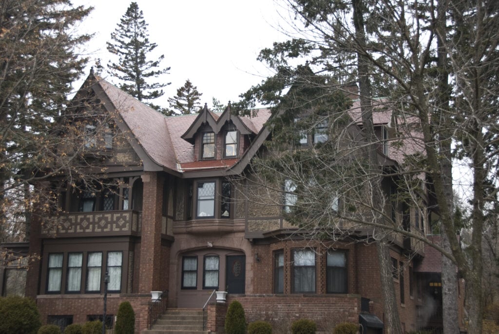 A large, historic, Tudor-style house with brick and timber details, multiple gables, tall windows, and a steeply pitched roof, surrounded by bare trees and evergreen pines on a cloudy day.