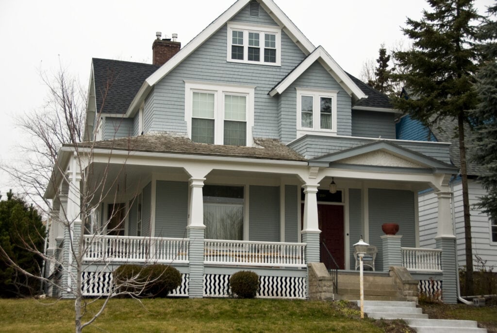 A blue-gray two-story house with white trim, a covered front porch with columns, and a red front door. Steps lead up to the porch, and there are bare trees and grass in the front yard.