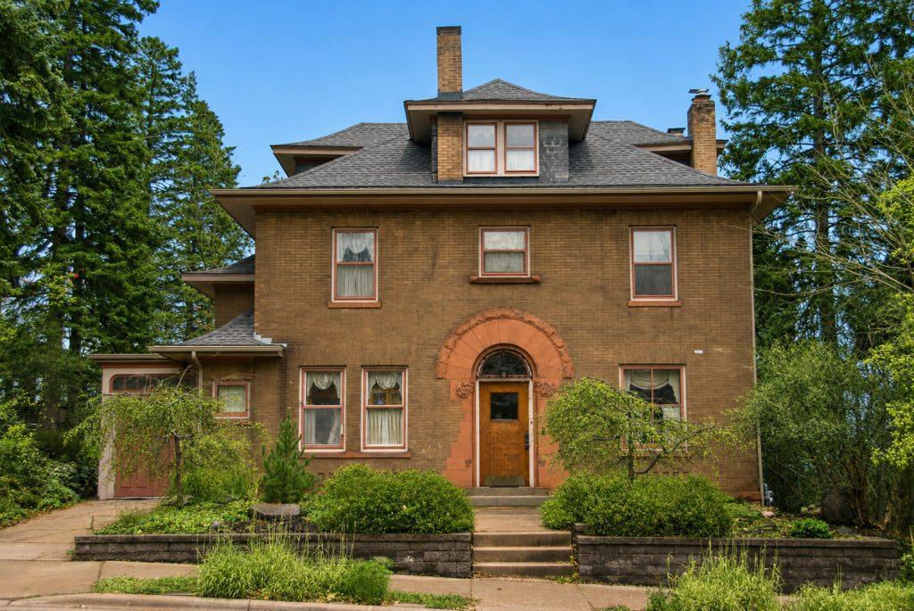A large, three-story brick house with a central arched doorway, multiple windows, a chimney, and surrounded by greenery and tall trees under a clear blue sky.