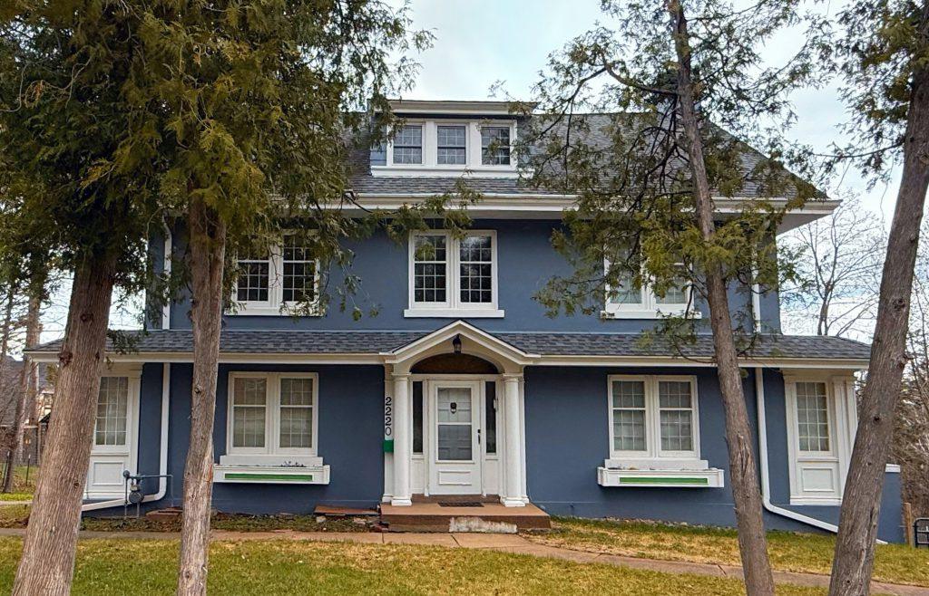 A two-story blue house with white trim, large windows, and a covered front entrance, surrounded by tall trees and grass on a cloudy day.