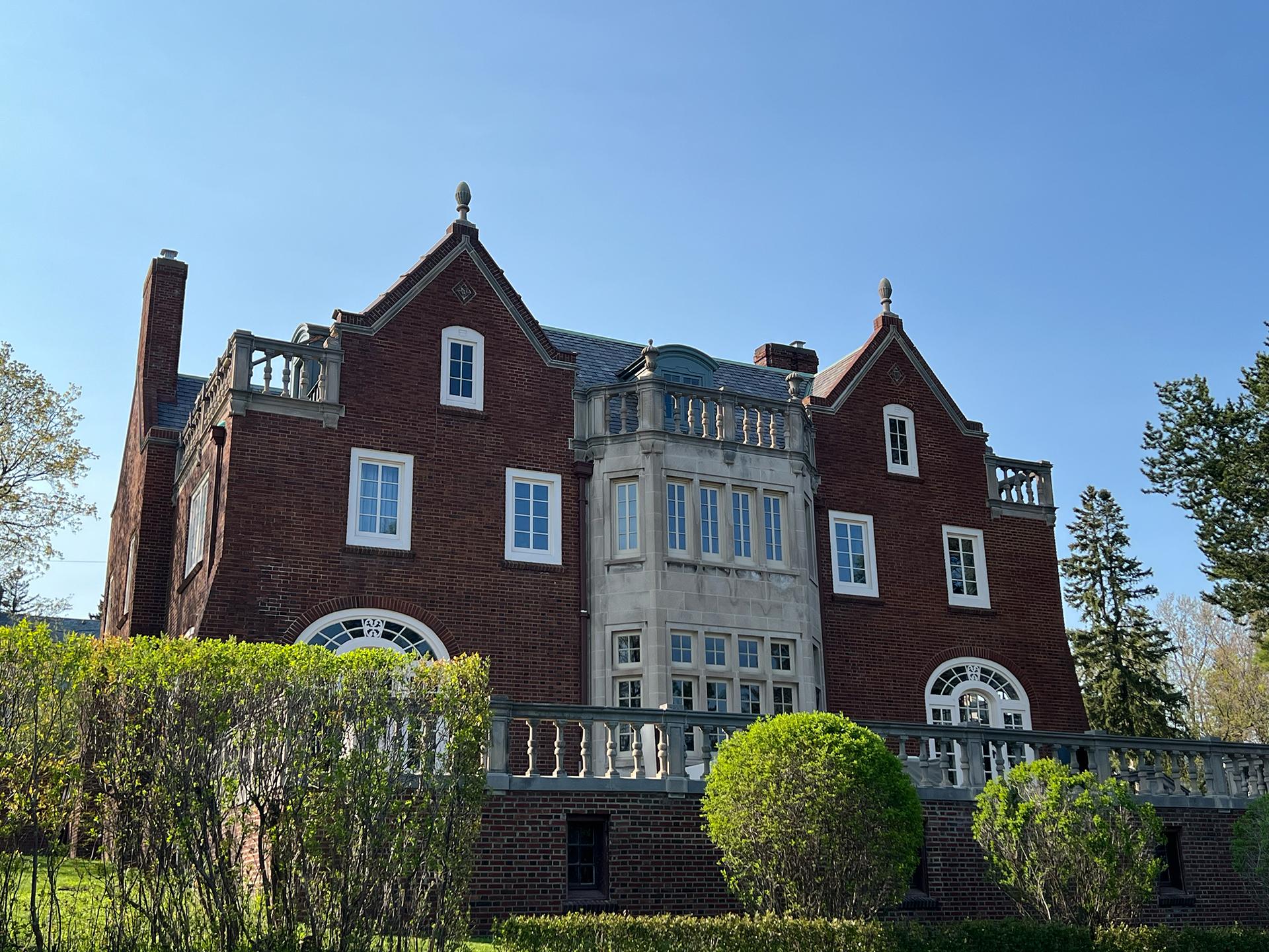 A large, red-brick mansion with white-trimmed windows, steep gabled roofs, and ornate stonework sits behind a manicured hedge on a clear, sunny day.