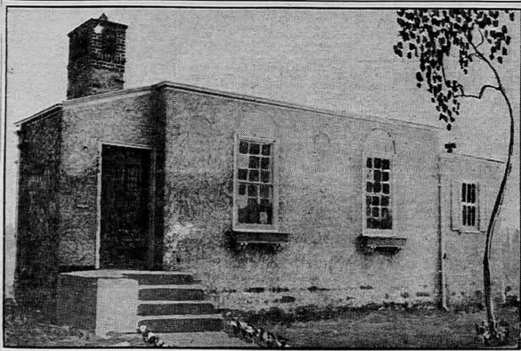 A black and white photo of a small, rectangular, single-story building with a chimney, two windows, and a door accessed by a short set of steps. A tree is visible on the right side of the image.