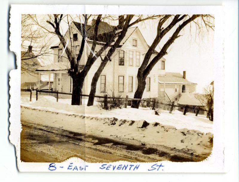 A sepia-toned photo of a large two-story house behind a fence and trees, with snow covering the ground and street. Handwritten text at the bottom reads “8 - East Seventh St.”.