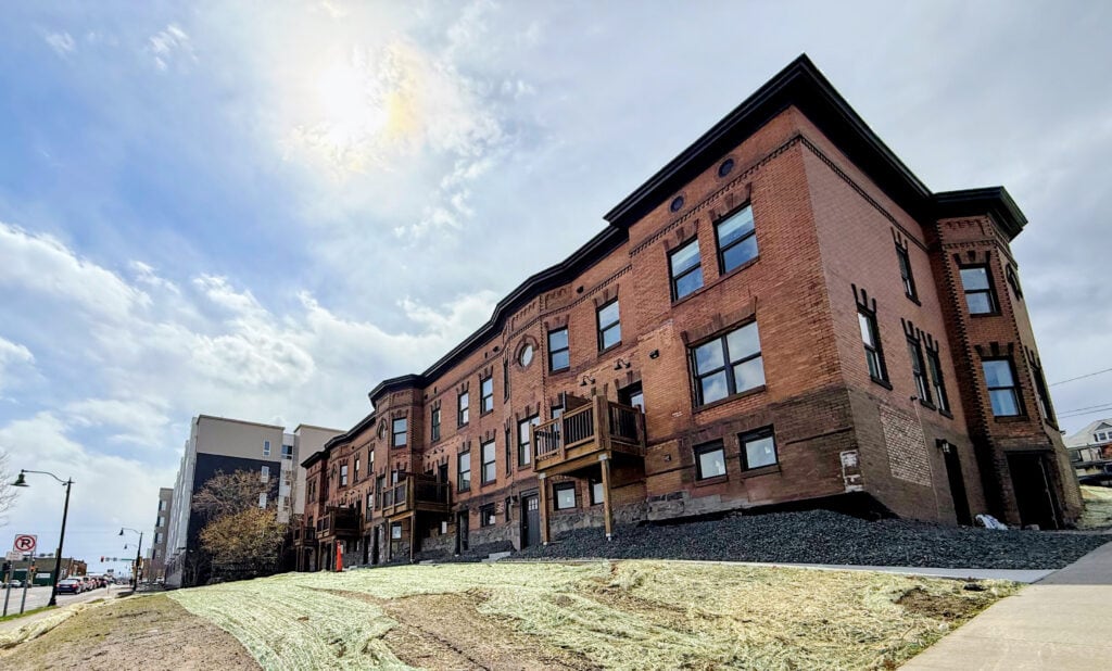 A row of historic red brick apartment buildings with small balconies sits on a sloped lawn under a bright, partly cloudy sky. Sidewalks and a city street are visible in the foreground.