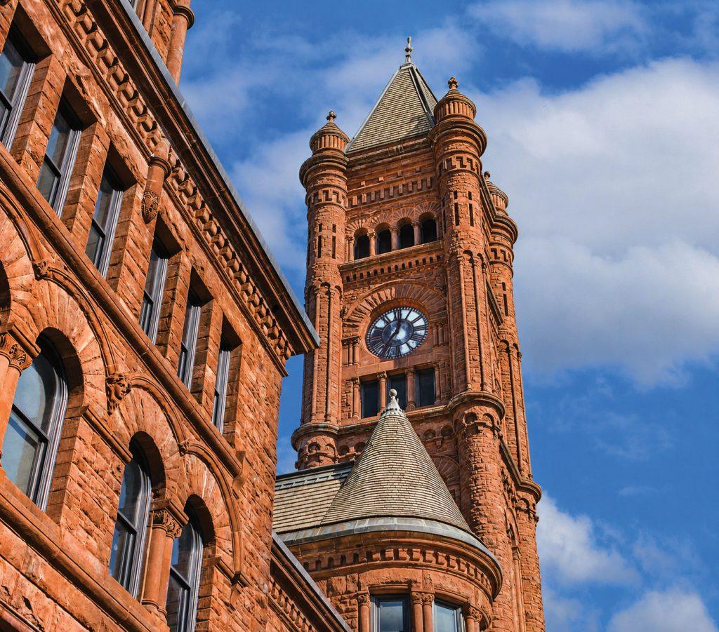 A tall, historic red brick clock tower with arched windows stands against a blue sky with scattered clouds. The architecture features detailed stonework and a steep, conical roof.