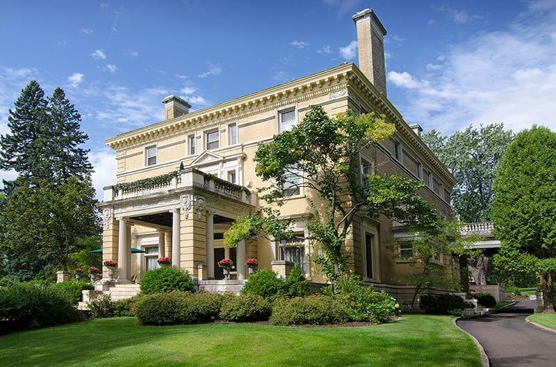 A large, elegant yellow mansion with white columns and trim, surrounded by green lawns, bushes, and trees under a blue sky with scattered clouds. Red flowers in planters accent the building.