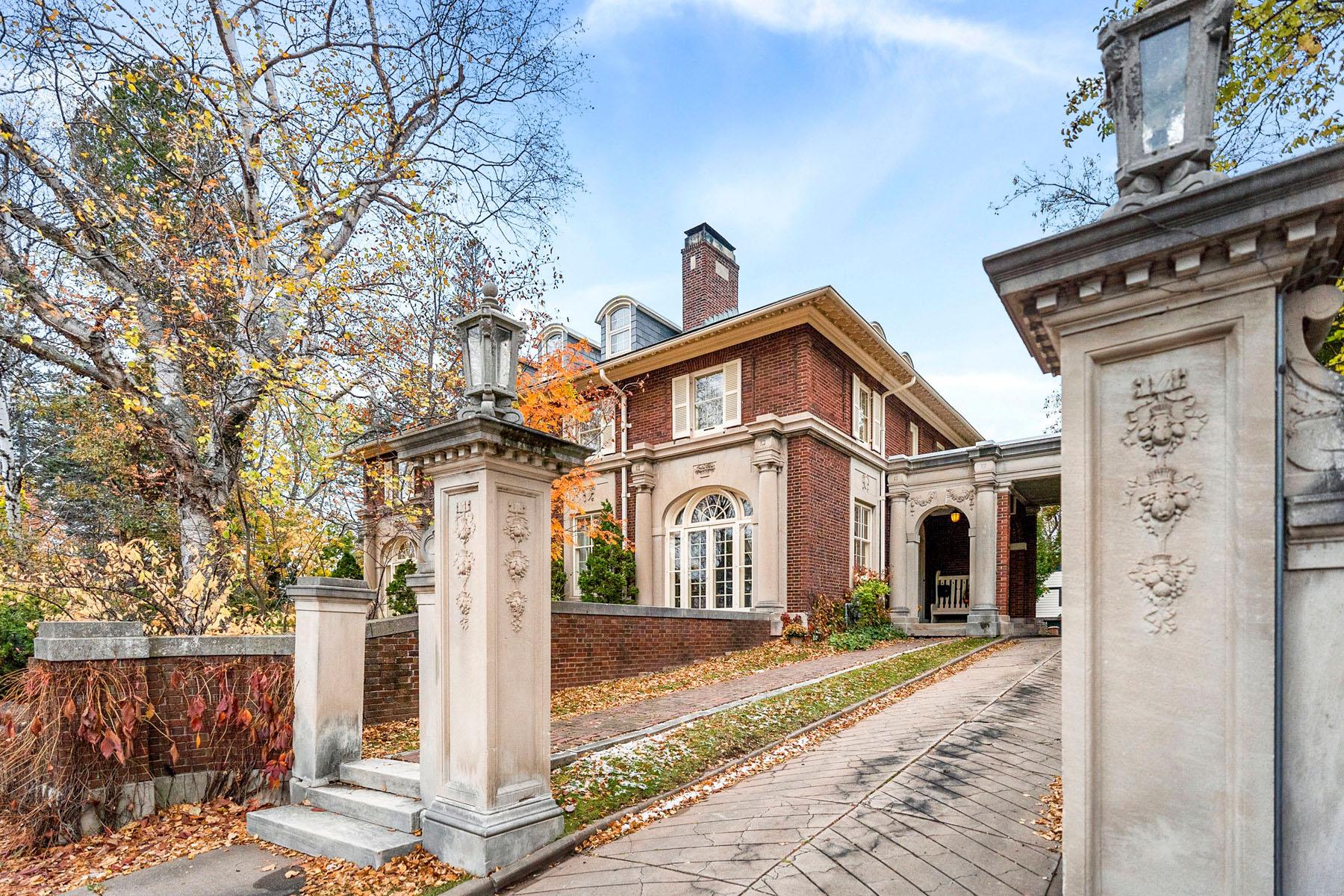 A large, elegant brick house with tall windows and a chimney, framed by ornate stone pillars and an iron gate, sits along a tree-lined driveway covered in autumn leaves.