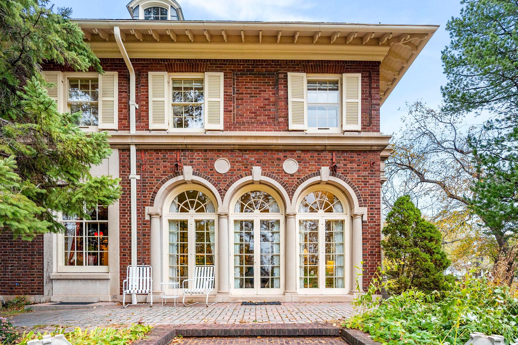 A historic brick house with large arched windows, white shutters, and a row of white patio chairs on a brick terrace, surrounded by greenery and trees.
